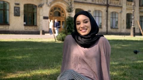 Young happy muslim girl in hijab is sitting on lawn in sun, watching at camera, builging on background, religious concept, relax concept.